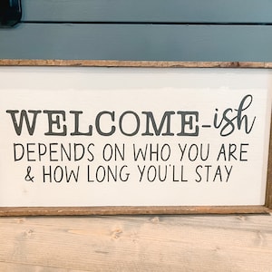 May include: A white wooden sign with a brown frame, featuring the text "WELCOME-ish DEPENDS ON WHO YOU ARE & HOW LONG YOU'LL STAY".