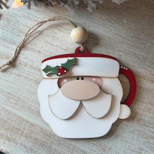 May include: A wooden Christmas bauble shaped like a Santa Claus mug. The bauble is painted white with red accents and features a green holly sprig with red berries.