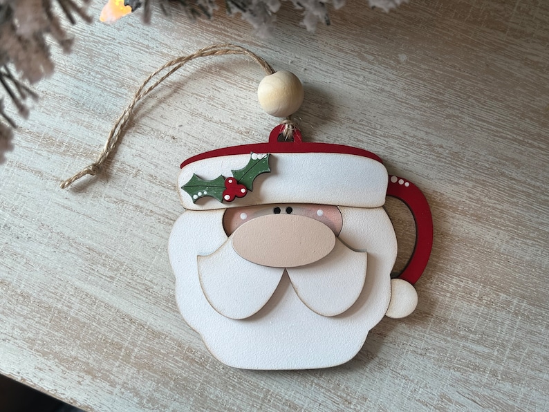 May include: A wooden Christmas bauble shaped like a Santa Claus mug. The bauble is painted white with red accents and features a green holly sprig with red berries.
