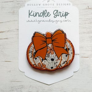 May include: A pumpkin-shaped Kindle Grip with an orange bow and floral design. The grip has a glittery orange border and the text "Kindle Grip" and "www.hgnovedesigns.com" on the backing card. The design is for fall.