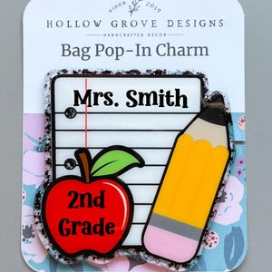 May include: A school-themed bag pop-in charm. It features a notepad with "Mrs. Smith", a red apple with "2nd Grade", and a yellow pencil. The charm is set against a floral pattern background.