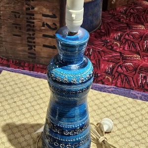 May include: A blue ceramic table lamp with a white lamp holder and a cream-coloured cord. The lamp has a textured surface with decorative patterns and a bulb socket. The lamp is approximately 30 cm tall.