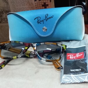May include: Ray-Ban sunglasses with a multi-coloured patterned frame, alongside a light blue case and a cleaning cloth. The case and cloth bear the Ray-Ban logo. The cloth has the text "GENUINE SINCE 1937". The sunglasses are on a white surface.