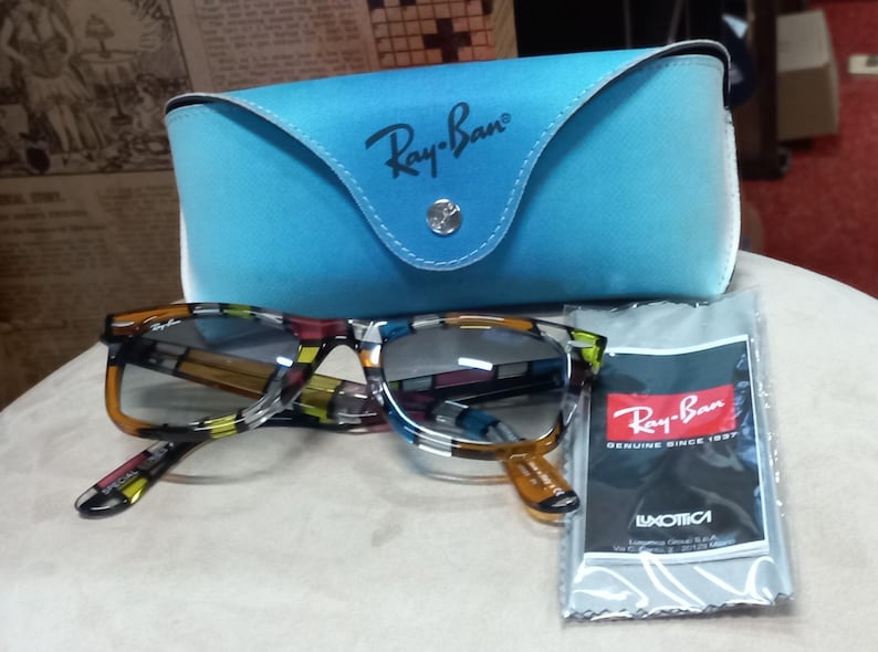 May include: Ray-Ban sunglasses with a multi-coloured patterned frame, alongside a light blue case and a cleaning cloth. The case and cloth bear the Ray-Ban logo. The cloth has the text "GENUINE SINCE 1937". The sunglasses are on a white surface.