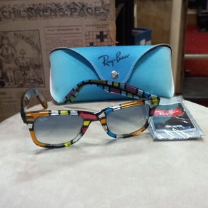 May include: A pair of Ray-Ban sunglasses with a vibrant, geometric design on the frames. They sit on a light surface, accompanied by a light blue Ray-Ban case and a cleaning cloth. The sunglasses feature gradient lenses, with the Ray-Ban logo clearly visible.