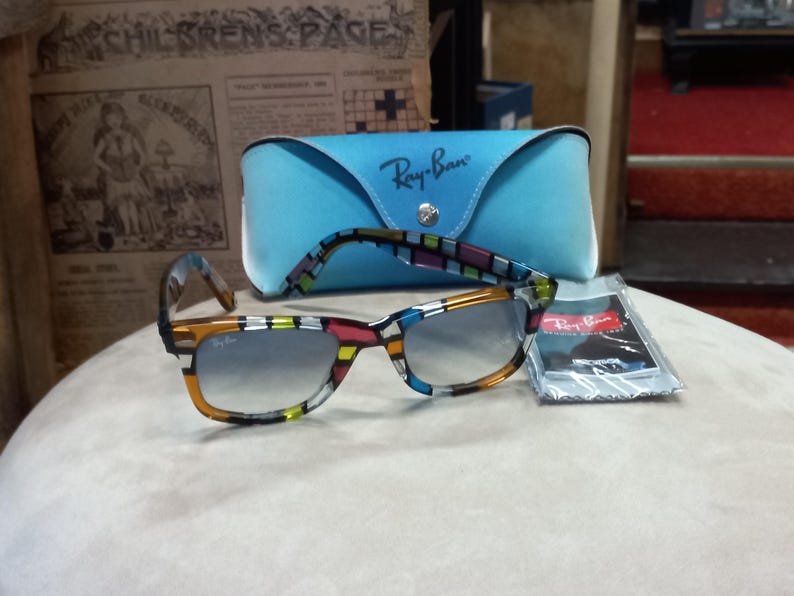 May include: A pair of Ray-Ban sunglasses with a vibrant, geometric design on the frames. They sit on a light surface, accompanied by a light blue Ray-Ban case and a cleaning cloth. The sunglasses feature gradient lenses, with the Ray-Ban logo clearly visible.
