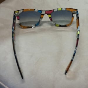 May include: Sunglasses with a colourful geometric pattern on the frames. The lenses have a gradient from blue to clear. The arms are black and brown with colourful accents. The sunglasses are set against a neutral background.