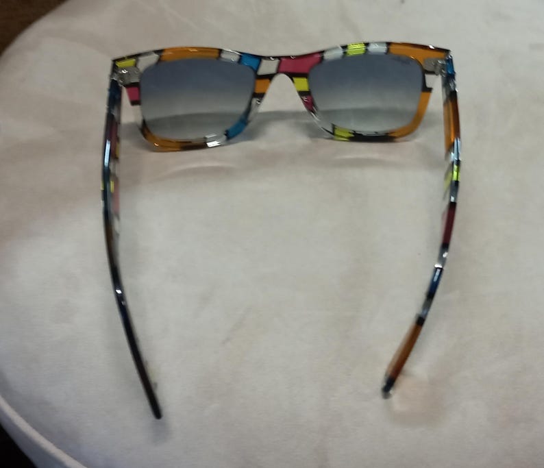 May include: Sunglasses with a colourful geometric pattern on the frames. The lenses have a gradient from blue to clear. The arms are black and brown with colourful accents. The sunglasses are set against a neutral background.