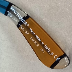May include: Close-up of the arm of Wayfarer sunglasses. The arm is brown and blue with the text "WAYFARER", "RB 2140", "1085/3F", "50022 2N", "Hand Made in Italy B CE".
