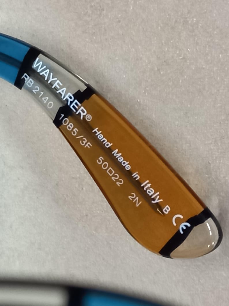 May include: Close-up of the arm of Wayfarer sunglasses. The arm is brown and blue with the text "WAYFARER", "RB 2140", "1085/3F", "50022 2N", "Hand Made in Italy B CE".