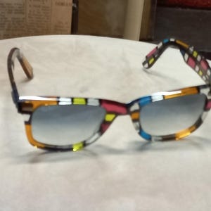 May include: Square-framed sunglasses with a vibrant mosaic pattern in orange, yellow, pink, and blue. The lenses have a gradient tint. The temples are patterned with a similar design. The sunglasses are a stylish accessory.