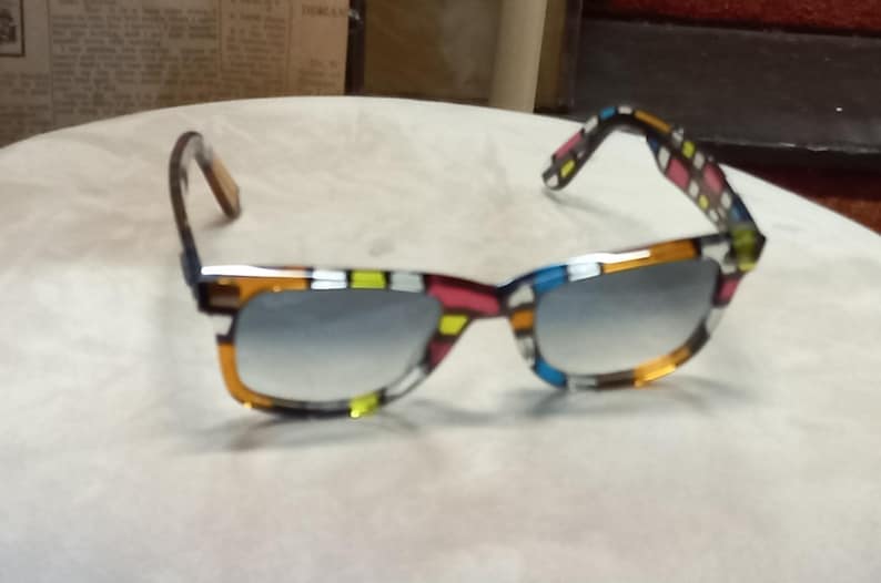 May include: Square-framed sunglasses with a vibrant mosaic pattern in orange, yellow, pink, and blue. The lenses have a gradient tint. The temples are patterned with a similar design. The sunglasses are a stylish accessory.