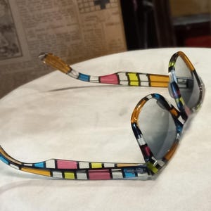 May include: Sunglasses with a vibrant geometric pattern in blue, pink, yellow, and orange. The frames feature a rectangular design, and the lenses are grey. The arms of the glasses are also patterned.