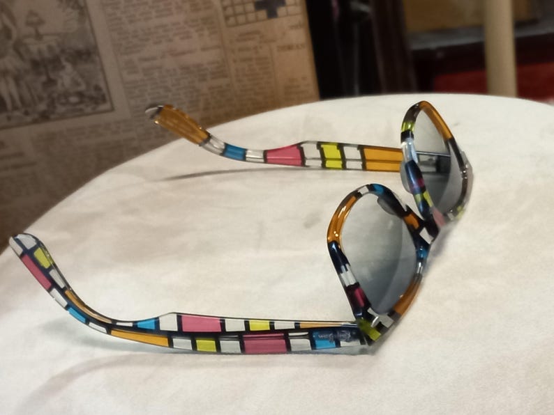 May include: Sunglasses with a vibrant geometric pattern in blue, pink, yellow, and orange. The frames feature a rectangular design, and the lenses are grey. The arms of the glasses are also patterned.