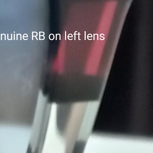 May include: Close-up of sunglasses with a dark frame and a red-tinted lens. The text "Genuine RB on left lens" is visible. The sunglasses are partially visible against a blurred background.