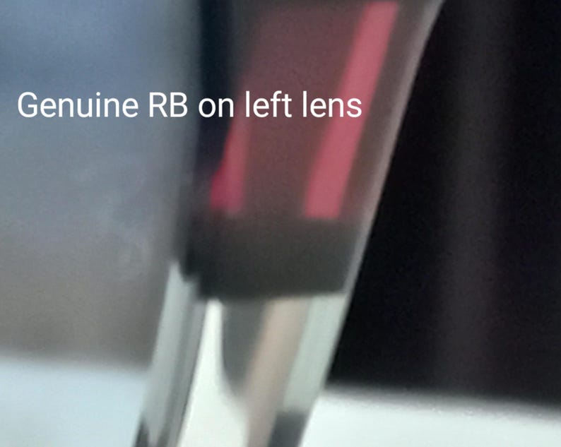 May include: Close-up of sunglasses with a dark frame and a red-tinted lens. The text "Genuine RB on left lens" is visible. The sunglasses are partially visible against a blurred background.