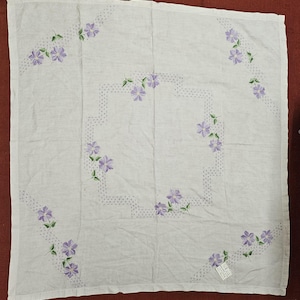 May include: A square white fabric cloth featuring embroidered purple flowers with green leaves arranged in a central rectangular pattern and along the four corners. The design includes small purple dotted lines connecting the floral motifs.