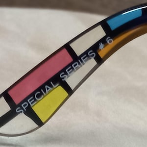 May include: Close-up of the arm of eyeglasses with a colourful geometric design. The design features rectangular blocks of pink, yellow, white, blue, and navy blue, separated by black lines. The text "SPECIAL SERIES #6" is printed on the arm.