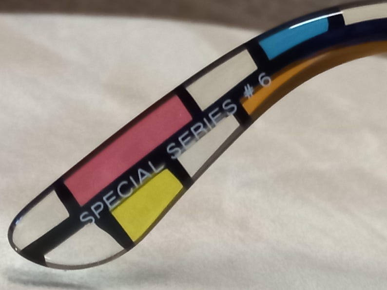 May include: Close-up of the arm of eyeglasses with a colourful geometric design. The design features rectangular blocks of pink, yellow, white, blue, and navy blue, separated by black lines. The text "SPECIAL SERIES #6" is printed on the arm.