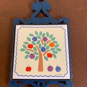 May include: A blue metal trivet with a square ceramic tile featuring a tree design with red apples, purple plums, blue and orange fruit. The tile is framed by a blue metal border with decorative flourishes.