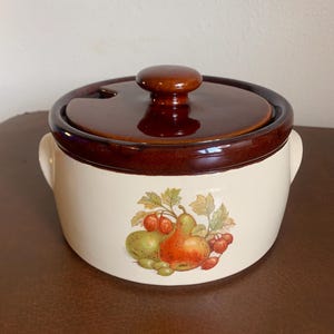 May include: A ceramic casserole dish with a lid. The dish is cream-colored with a brown rim and lid. A fruit design of pears, tomatoes, and berries decorates the front. The lid has a small knob handle. The dish has two small side handles.