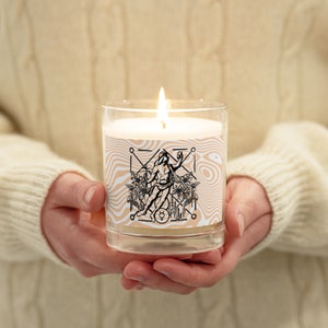 May include: A white candle with a black and white design of a figure standing in a circle with geometric shapes and plants. The candle is in a clear glass jar.