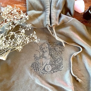May include: A green hooded sweatshirt with a black and white graphic print of a woman with long hair and a rose. The graphic also includes a symbol that looks like a circle with a cross inside.