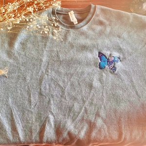 Könnte beinhalten: Ein grauer Pullover mit einem blauen Schmetterling, der auf der Vorderseite gestickt ist. Der Schmetterling hat lila und schwarze Akzente.