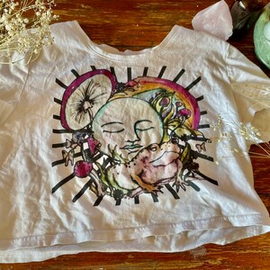 Puede incluir: Camiseta blanca recortada con un estampado gráfico colorido de una luna, una rana, hongos y un arcoíris. El gráfico está rodeado de rayas blancas y negras.