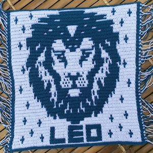 May include: A crocheted square with a blue lion design and the word "LEO" in white. The square has a white fringe border.