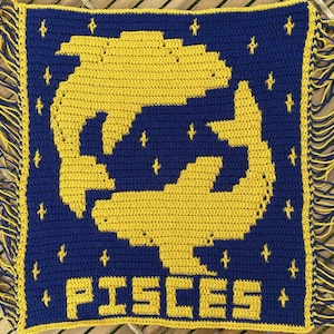 May include: A crocheted square with a blue background and yellow fish design. The word "PISCES" is written in yellow letters at the bottom of the square.