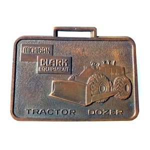 May include: A bronze-coloured metal keychain featuring a tractor dozer with a blade. The keychain has the text "MICHIGAN CLARK EQUIPMENT TRACTOR DOZER" on it.