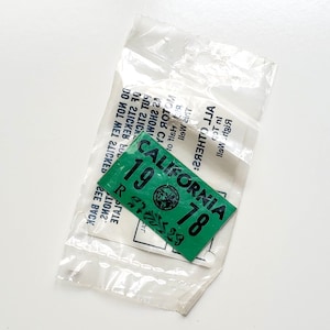May include: A green California 1978 license plate sticker in a clear plastic bag. The sticker has black text and a state seal. The bag has text printed on it.