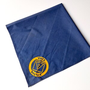 May include: A blue fabric neckerchief with a yellow circle containing the text "YMCA" and a triangle with a cross inside. The text "Ragger" is below the circle.