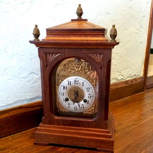 May include: A wooden mantel clock with a dark brown finish, featuring a square base, a pediment top with three brass finials, and an arched glass front. The clock face is white with black Arabic numerals and ornate gold-colored metal filigree.