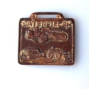 May include: A rectangular, brown metal pendant with a handle at the top. The word "CATERPILLAR" is at the top. Below are embossed images of construction vehicles. The pendant is likely a promotional item or collectible.