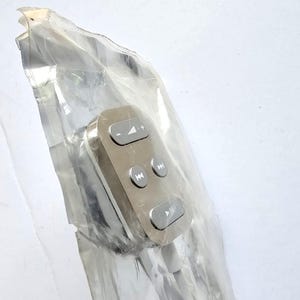 May include: A silver-toned remote control unit with three grey buttons featuring white icons for volume adjustment, track skipping, and play-pause functions, encased in a clear, crinkled plastic protective sleeve with a white cable attached.