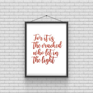 May include: A framed print with a white background and black frame. The print features red glitter lettering that reads "For it is the cracked who let in the light."