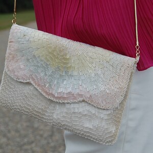 Puede incluir: Un bolso de mano blanco de cuentas con una correa de cadena dorada. El bolso tiene un borde festoneado y un diseño iridiscente.