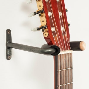 Peut inclure: Un support mural en métal noir pour guitare avec un bouton en bois, tenant une guitare acoustique brune.