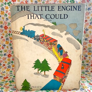 Könnte beinhalten: Ein Vintage-Kinderbuchcover mit einer farbenfrohen Illustration einer Lokomotive, die eine Reihe von Waggons einen schneebedeckten Hügel hinaufzieht. Der Buchtitel lautet "The Little Engine That Could" mit den Worten "Trade Mark" unter dem Titel.