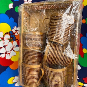 May include: A set of woven rattan cups and saucers with handles, packaged in a clear plastic wrap. The cups and saucers are a light brown color and have a textured, ribbed design. The set is contained within a matching woven tray.
