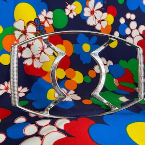 May include: A silver metal hand grip exercise tool with a curved design. The tool is photographed on a colorful fabric background with a floral pattern.