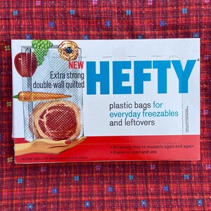 Puede incluir: Una caja de bolsas de plástico Hefty para congelar y guardar sobras. La caja es roja y blanca y presenta una foto de una bolsa con un trozo de carne dentro. El texto en la caja dice "Nuevo Extra fuerte acolchado de doble pared" y "bolsas de plástico para congelar todos los días y sobras".