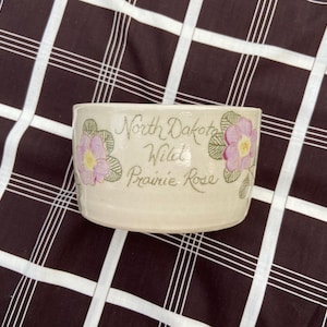 Könnte beinhalten: Eine weiße Keramikschüssel mit einem Blumendesign und dem Text "North Dakota Wild Prairie Rose".