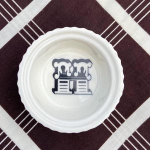 May include: A white ceramic bowl with a black and white graphic of a house with the letter "M" on the front. The bowl is on a brown and white checkered tablecloth.