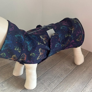 Könnte beinhalten: Ein marineblauer Hundemantel mit einem bunten Schmetterlingsmuster. Der Mantel hat einen Reißverschluss und eine kleine Tasche. Die Schmetterlinge sind in verschiedenen Farben umrandet und schaffen ein lebendiges Design für Tierbekleidung.