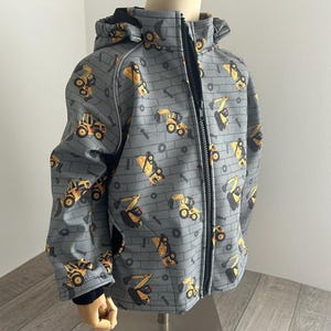 Könnte beinhalten: Graue Kapuzenjacke mit schwarzem Reißverschluss und einem Muster aus gelben und schwarzen Baumaschinen. Die Jacke hat ein ziegelsteinartiges Hintergrundmuster und einen schwarzen Kragen.