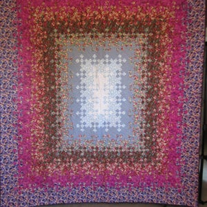 Könnte beinhalten: Eine Steppdecke mit einem großen, komplizierten Muster aus kleinen Blumen in Rosa-, Lila- und Brauntönen. Die Mitte der Steppdecke ist ein Quadrat aus hellblauem Stoff mit einem Rautenmuster.