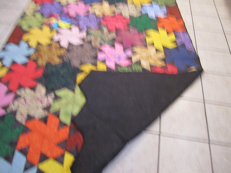 May include: A colourful patchwork quilt featuring a range of geometric shapes and patterns. The quilt is made from different fabrics in shades of red, green, blue, yellow and pink. The quilt is folded in half and the underside is black.